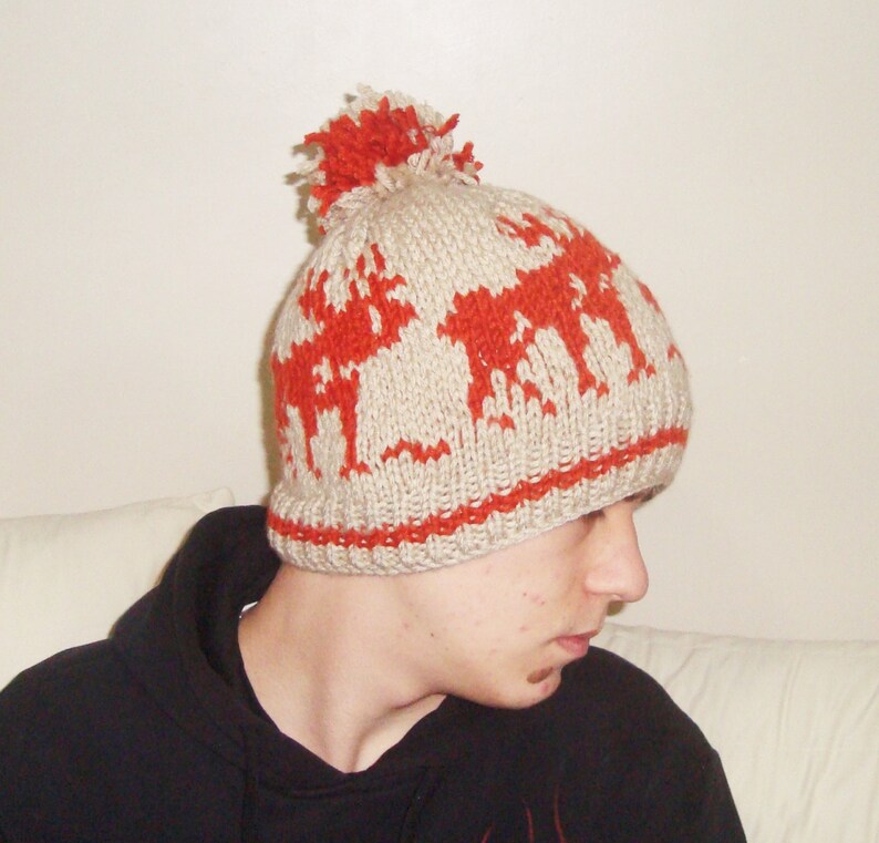 May include: A beige knitted hat featuring a red reindeer design and a red pom-pom. The hat has a red band at the bottom. This winter hat is made of a soft, warm material, ideal for cold weather.
