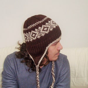 May include: A brown knit hat with a white geometric pattern. The hat has ear flaps and a long tassel.