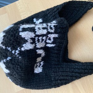 May include: A black and white knitted hat with a long strap. The hat features a white snowflake pattern on the front. The strap is made of a thick, twisted black yarn. The hat is likely handmade.