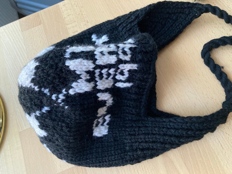 May include: A black and white knitted hat with a long strap. The hat features a white snowflake pattern on the front. The strap is made of a thick, twisted black yarn. The hat is likely handmade.