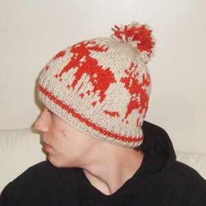 May include: A beige knitted hat featuring a red pattern and a matching pom-pom. The hat has a red band around the bottom. The hat is made of a soft, warm material, ideal for cold weather.