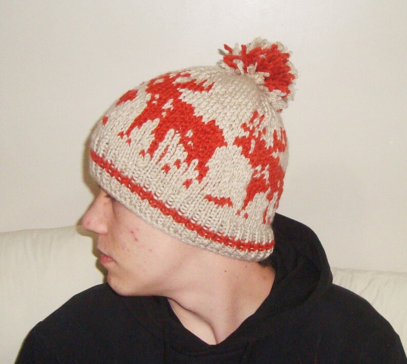 May include: A beige knitted hat featuring a red pattern and a matching pom-pom. The hat has a red band around the bottom. The hat is made of a soft, warm material, ideal for cold weather.