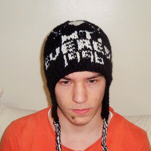 May include: A black knit hat with white lettering and ear flaps. The hat has a repeating pattern of white text. The ear flaps are braided with black and white yarn. The person in the image is wearing an orange shirt.