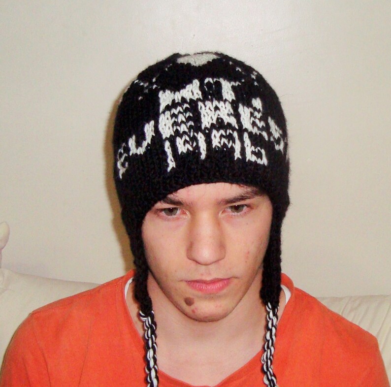 May include: A black knit hat with white lettering and ear flaps. The hat has a repeating pattern of white text. The ear flaps are braided with black and white yarn. The person in the image is wearing an orange shirt.
