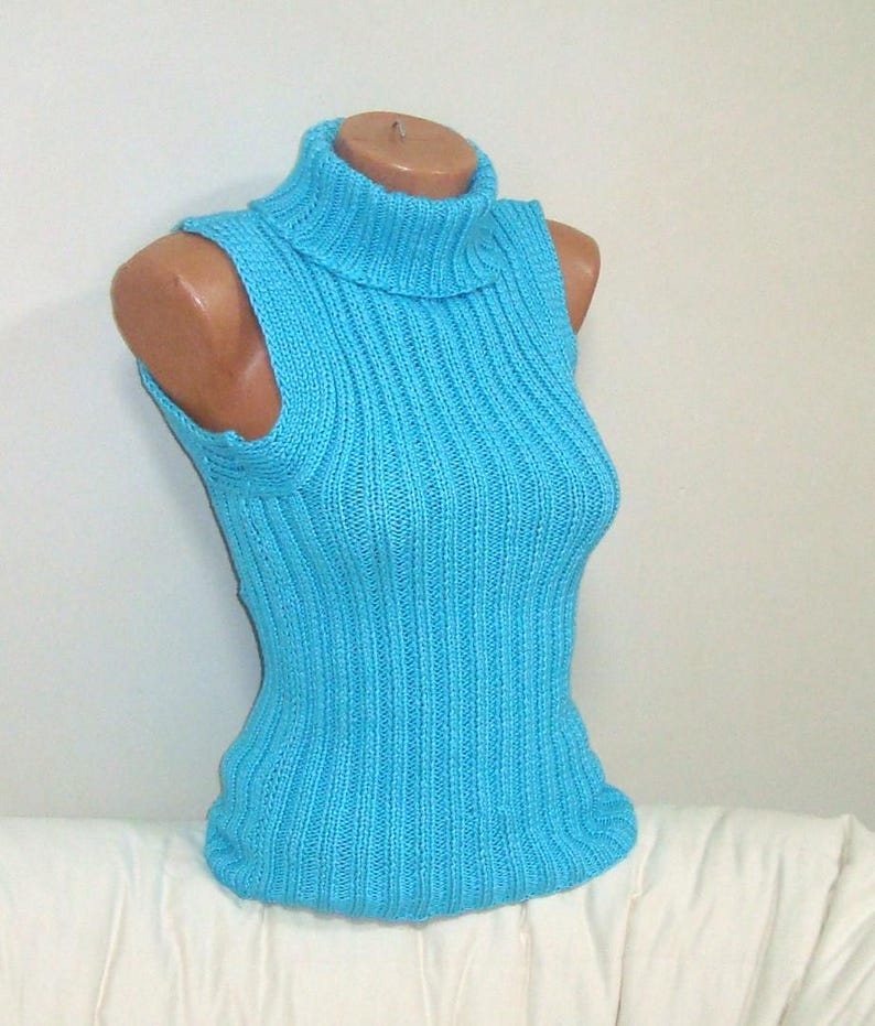 May include: A sleeveless, light blue knitted turtleneck top. The top is displayed on a mannequin and features a ribbed texture. The turtleneck is folded over. The top is made of a soft, possibly wool, material.