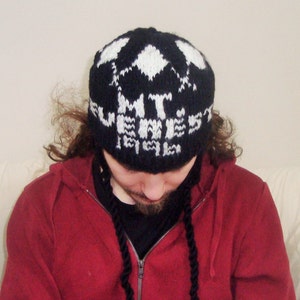 May include: A person wearing a black and white knit beanie with the words "MT. EVEREST" on it. The beanie has long black cords hanging down the front. The person is wearing a red hoodie.