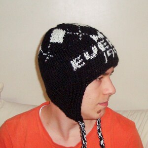May include: A black and white knit hat with ear flaps. The hat features a geometric pattern in white against a black background. The ear flaps have braided black and white cords.
