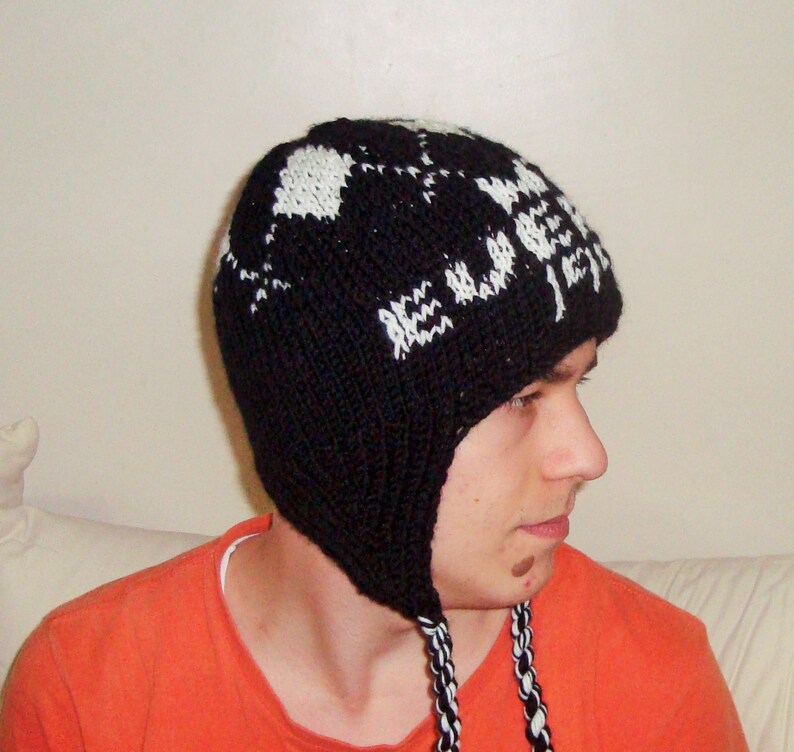 May include: A black and white knit hat with ear flaps. The hat features a geometric pattern in white against a black background. The ear flaps have braided black and white cords.