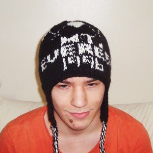 May include: Black knit hat with white text that reads "MUTATE". The hat has ear flaps with black and white braided cords. The person in the image is wearing an orange shirt.