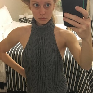 K&ouml;nnte beinhalten: Grauer Strickpullover mit Rollkragen und Zopfmuster, der einen Ausschnitt im R&uuml;cken hat. Der Pullover wird &uuml;ber einer schwarzen Hose getragen.