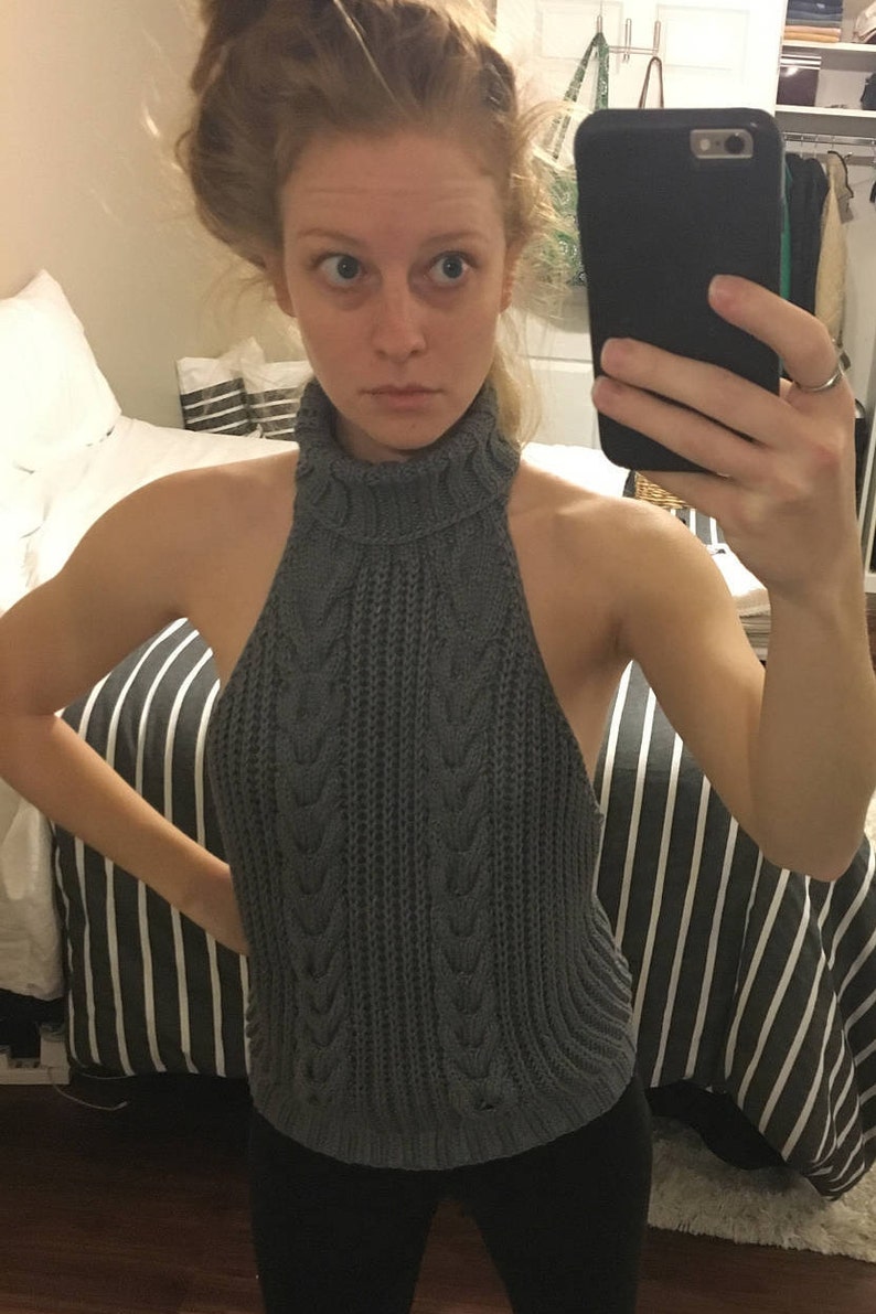 K&ouml;nnte beinhalten: Grauer Strickpullover mit Rollkragen und Zopfmuster, der einen Ausschnitt im R&uuml;cken hat. Der Pullover wird &uuml;ber einer schwarzen Hose getragen.