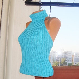 May include: A turquoise halter top with a ribbed knit design. The top features a turtleneck and a fitted silhouette. The garment is displayed on a mannequin, showcasing its shape and texture. The background includes a glimpse of a cityscape.
