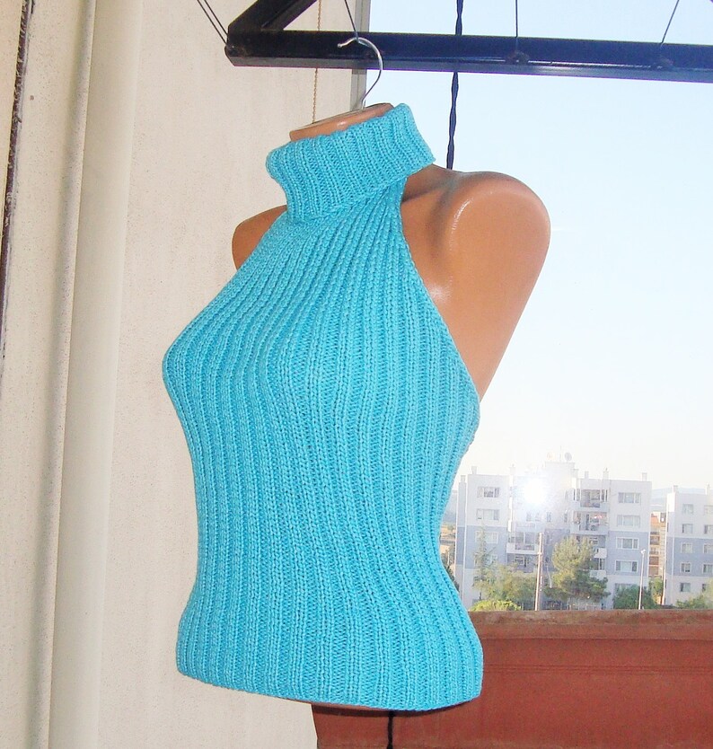 May include: A turquoise halter top with a ribbed knit design. The top features a turtleneck and a fitted silhouette. The garment is displayed on a mannequin, showcasing its shape and texture. The background includes a glimpse of a cityscape.