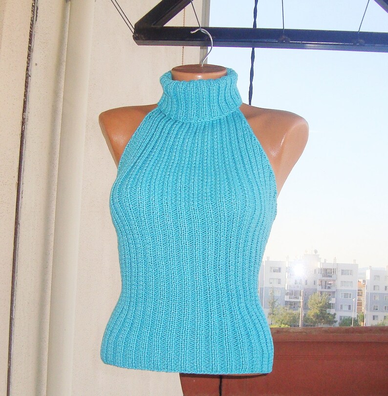 May include: A turquoise halter top with a ribbed knit design. The top features a high turtleneck and a fitted silhouette. The garment is displayed on a mannequin, showcasing its shape and texture. The background includes a view of a cityscape.