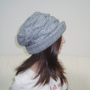 May include: A gray knitted beanie with a cable knit pattern. The beanie has a slouchy fit and a wide brim.