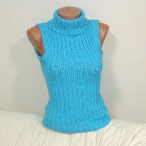 May include: A sleeveless, turquoise blue turtleneck sweater. The ribbed knit top is displayed on a mannequin. The sweater has a high, folded collar and a fitted silhouette. The garment is made of a soft, possibly wool, material.