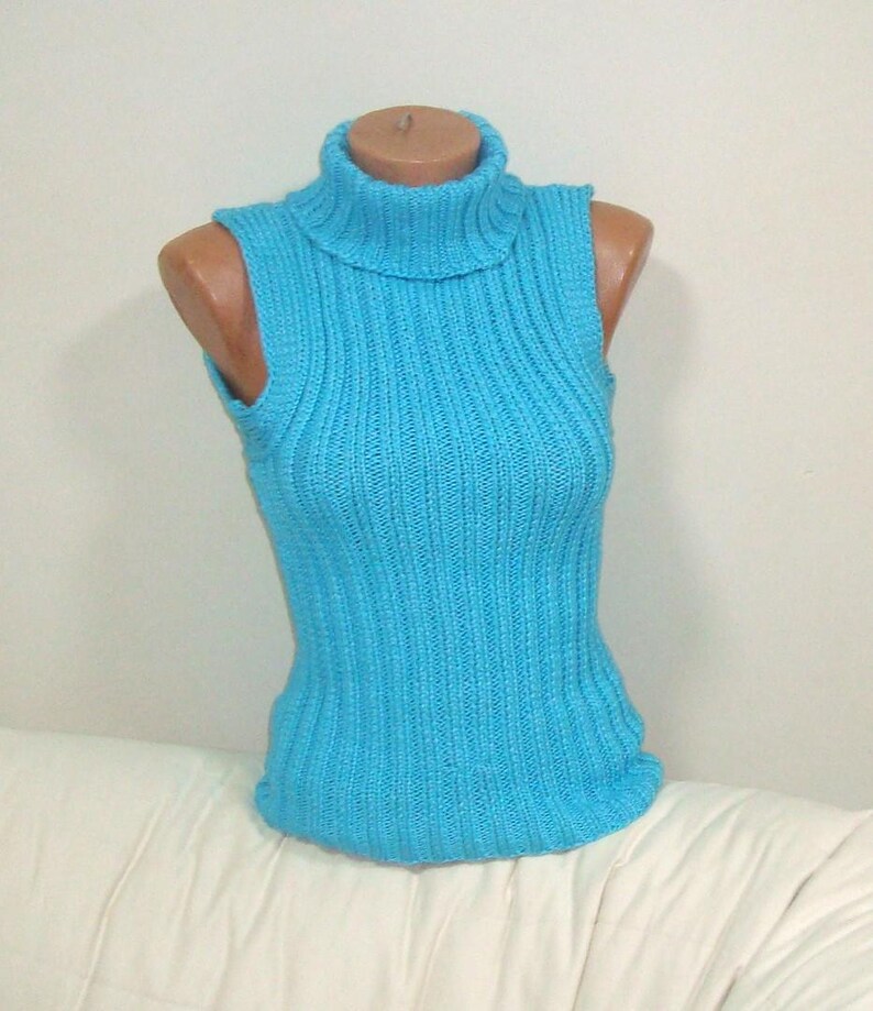 May include: A sleeveless, turquoise blue turtleneck sweater. The ribbed knit top is displayed on a mannequin. The sweater has a high, folded collar and a fitted silhouette. The garment is made of a soft, possibly wool, material.
