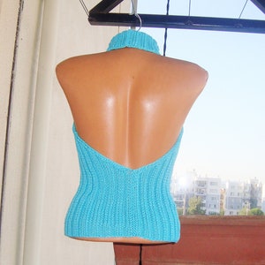 May include: A turquoise halter top with a ribbed knit design. The top features a high, ribbed collar and a low back. The garment is displayed on a mannequin, hanging from a clothesline against a bright sky.