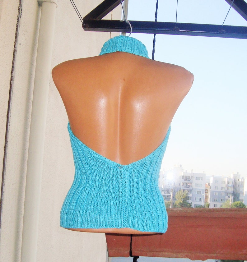May include: A turquoise halter top with a ribbed knit design. The top features a high, ribbed collar and a low back. The garment is displayed on a mannequin, hanging from a clothesline against a bright sky.