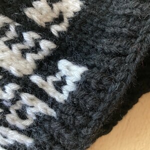 May include: Close-up of a knitted item with a black background and a white pattern. The texture of the yarn is visible, with the white pattern creating a contrast against the dark backdrop. The item appears to be handmade.