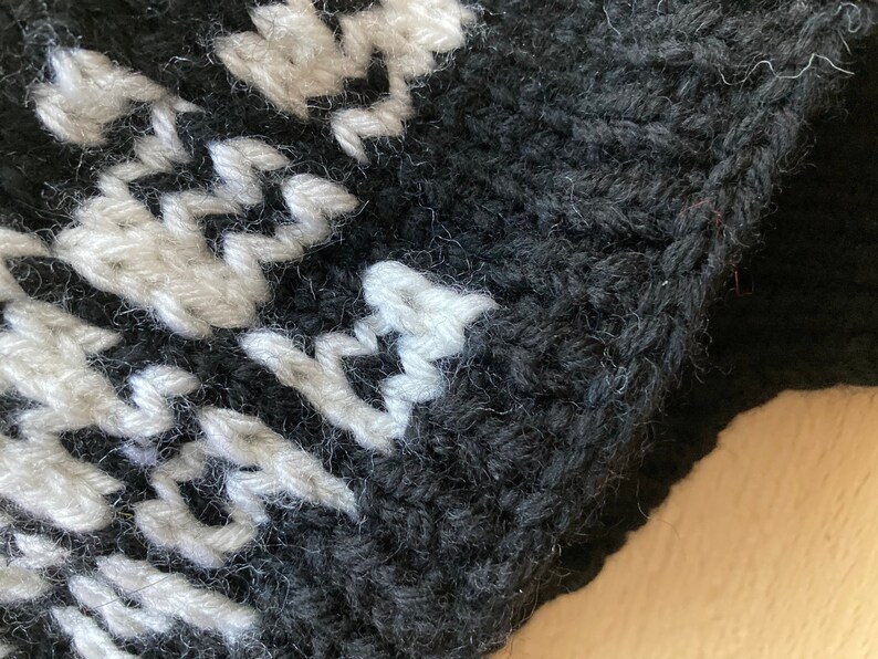 May include: Close-up of a knitted item with a black background and a white pattern. The texture of the yarn is visible, with the white pattern creating a contrast against the dark backdrop. The item appears to be handmade.
