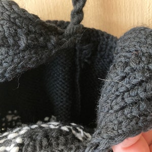 May include: Close-up of a black knitted bag with a textured, woven design. The bag's opening is visible, revealing a black interior and a white and black patterned base. A black handle is at the top.