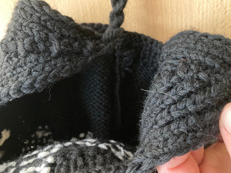 May include: Close-up of a black knitted bag with a textured, woven design. The bag's opening is visible, revealing a black interior and a white and black patterned base. A black handle is at the top.