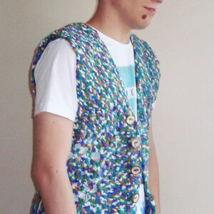 May include: A person wearing a white t-shirt with a blue and green crocheted vest with fringe. The vest has wooden buttons.