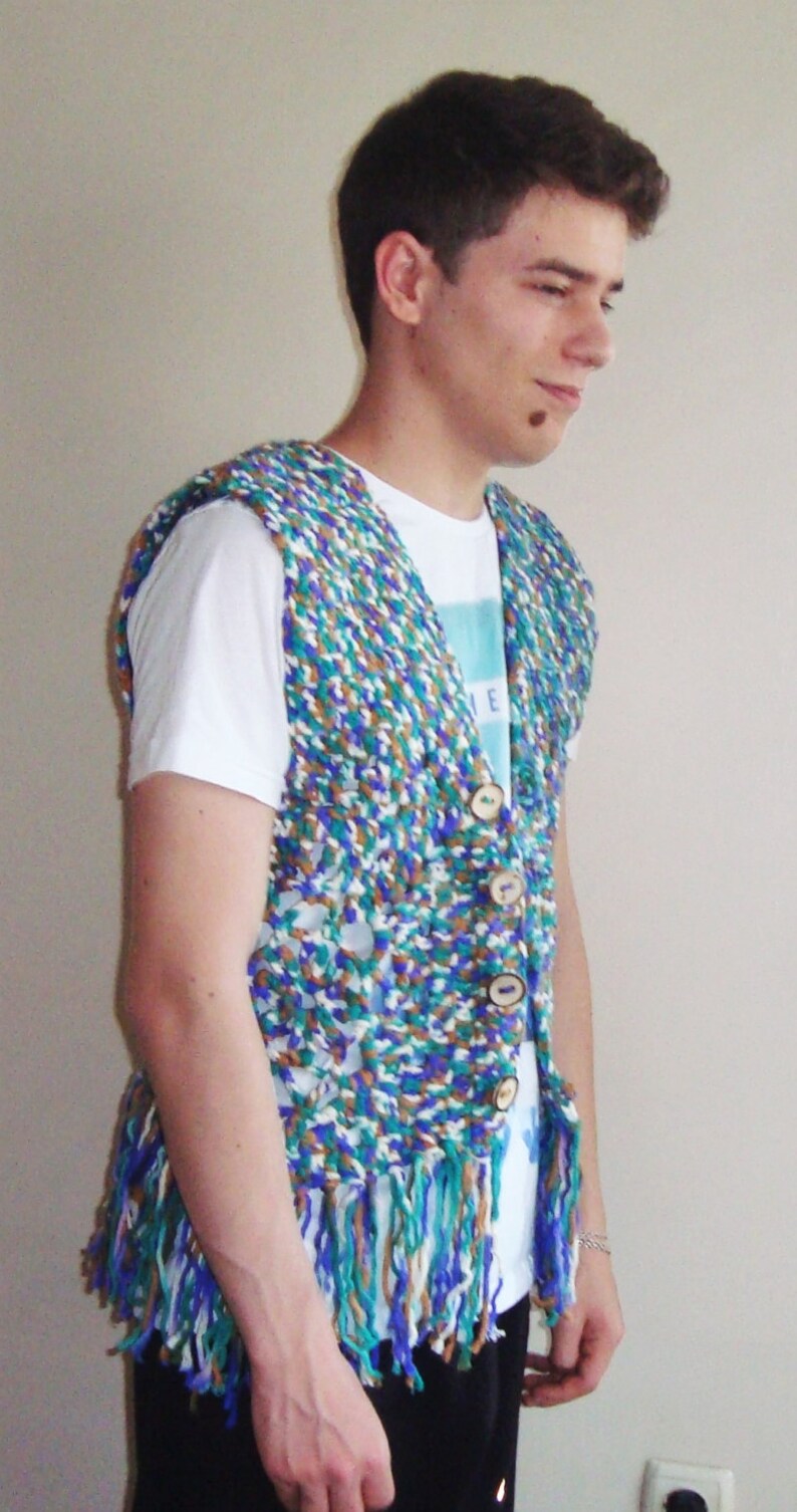 May include: A person wearing a white t-shirt with a blue and green crocheted vest with fringe. The vest has wooden buttons.