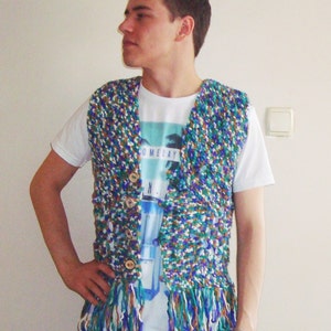 May include: A person wearing a multi-colored crocheted vest with fringe. The vest has a blue, green, white, and brown color scheme. The person is wearing a white t-shirt with the text "Someday" printed on it.