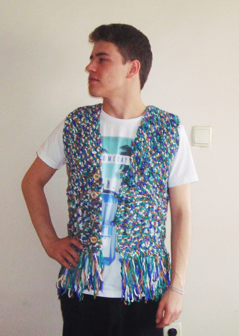 May include: A person wearing a multi-colored crocheted vest with fringe. The vest has a blue, green, white, and brown color scheme. The person is wearing a white t-shirt with the text "Someday" printed on it.