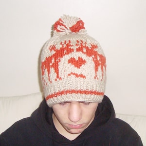 May include: A beige and orange knitted hat with a pom-pom. The hat has a repeating pattern of orange reindeer and hearts. The hat is made of a soft, textured yarn, suitable for cold weather.