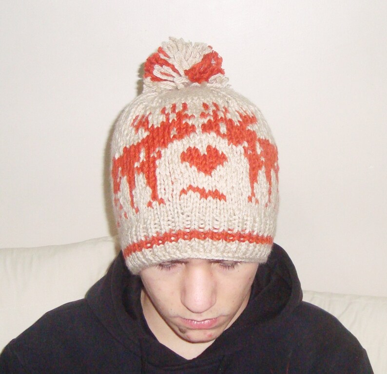 May include: A beige and orange knitted hat with a pom-pom. The hat has a repeating pattern of orange reindeer and hearts. The hat is made of a soft, textured yarn, suitable for cold weather.