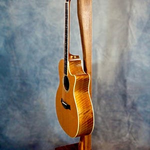 May include: A wooden guitar stand with a natural finish, holding an acoustic guitar with a brown wood body and a natural wood neck. The guitar has a light brown wood grain pattern.