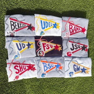 May include: A collection of nine sweatshirts in white, gray, and black, each featuring a decorative pennant applique with university acronyms including BERKLEE, UDEL, BAMA, UD, FSU, JSU, SHU, BSU, and GW, accented with stars, ribbons, and mascot logos.