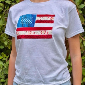 May include: White t-shirt featuring a fabric American flag design. The flag has a blue field with white stars, red and white stripes, and floral patterned stripes. The shirt is a classic crew neck style.