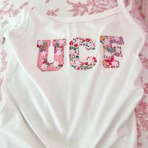 May include: White ribbed vest top with the letters "UCF" appliquéd across the front. The letters are made of floral fabric in shades of pink, red, and green. The vest top has thin straps.