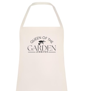 Puede incluir: Delantal color crema con cuello halter. El delantal presenta el texto "QUEEN OF THE GARDEN" en negro, con una ilustración de carretilla y el nombre "JENNIFER" debajo. Adecuado para jardinería.