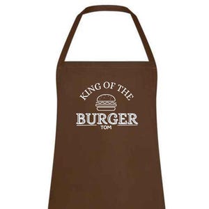 May include: A brown apron with a neck loop and waist ties. The apron features the text "KING OF THE BURGER" above a burger graphic, with "TOM" below. The text and graphic are in white.