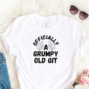 Può includere: T-shirt bianco con la scritta "OFFICIALLY A GRUMPY OLD GIT" in nero. La maglietta è abbinata a jeans blu, scarpe da ginnastica bianche, occhiali da sole e un bouquet di fiori rosa e bianchi.