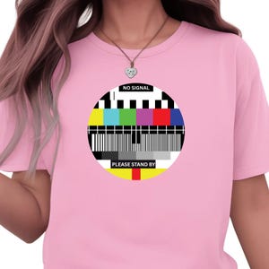 May include: A pale pink t-shirt with a circular television test pattern design. The graphic includes the words "NO SIGNAL" and "PLEASE STAND BY". The shirt has a crew neck and short sleeves.