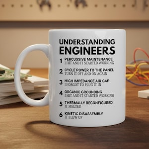 May include: White ceramic mug featuring the text "UNDERSTANDING ENGINEERS" and a list of troubleshooting steps. The mug has a curved handle and is on a wooden surface. The text includes phrases like "Percussive Maintenance" and "Kinetic Disassembly".