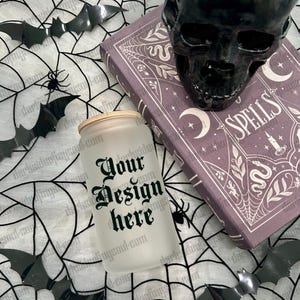 May include: A frosted glass tumbler with the text "Your Design Here" in black gothic lettering. A black skull and a purple book with the word "SPELLS" are in the background. Black bat and spider decorations are scattered around.