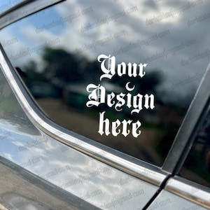 May include: A white vinyl decal with the text "Your Design here" in a gothic font. The decal is applied to the back window of a silver car.