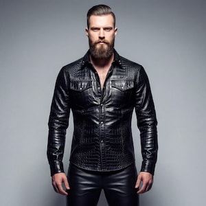 Puede incluir: Una persona con una camisa de cuero negra y pantalones a juego, posando sobre un fondo gris. La camisa tiene una textura y dos bolsillos en el pecho. Los pantalones son ajustados y la persona lleva zapatos negros. El conjunto es elegante.