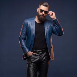 K&ouml;nnte beinhalten: Eine Person tr&auml;gt einen blauen Lederblazer, ein schwarzes T-Shirt, eine schwarze Lederhose und eine schwarze Sonnenbrille. Die Person steht mit einer Hand in der Tasche und der anderen an der Sonnenbrille. Der Hintergrund ist dunkelblau.
