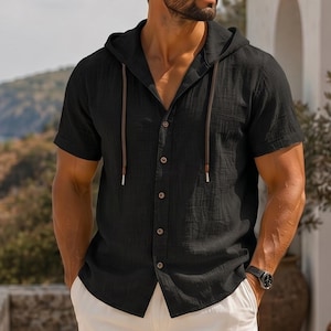 Pode incluir: Camisa preta com capuz de manga curta com botões. A camisa tem uma aparência texturizada e cordões marrons. O relógio é preto. A camisa é de estilo casual.