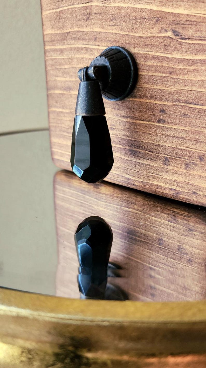 Peut inclure: Poign&eacute;e de cabinet en cristal noir avec une finition nickel bross&eacute;. La poign&eacute;e est mont&eacute;e sur une porte de cabinet en bois.