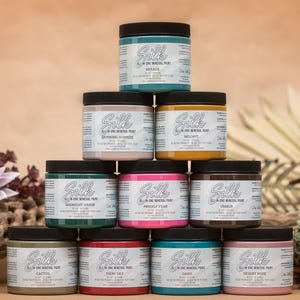 May include: A stack of ten 8 oz. jars of Silk All-in-One Mineral Paint in various colors, including Midnight Green, Prickly Pear, Lavender, and Desert Rose. The jars are arranged in a pyramid shape on a brown surface.