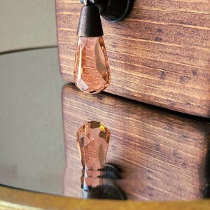 Peut inclure: Gros plan d'une bo&icirc;te en bois avec un bouton en cristal. Le bouton est en forme de larme et est de couleur p&ecirc;che clair. La bo&icirc;te est de couleur brun fonc&eacute; et pr&eacute;sente un motif de grain de bois.